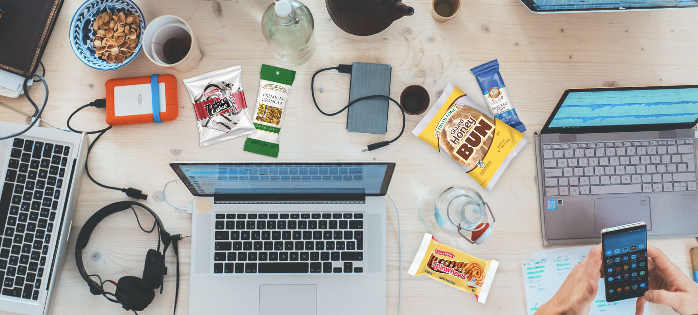 A top view of laptops and other electronic items on a desk at a college or university. Snacks are scattered around  the desk. The college snacks include, pecan pinwheels, lemon doodles, honey buns, zebra cakes, and premium granola. 