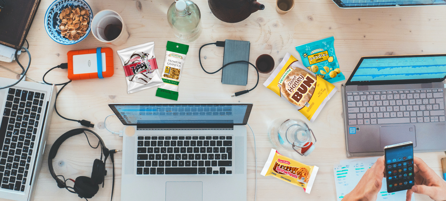 A top view of laptops and other electronic items on a desk at a college or university. Snacks are scattered around  the desk. The college snacks include, pecan pinwheels, lemon doodles, honey buns, zebra cakes, and premium granola. 