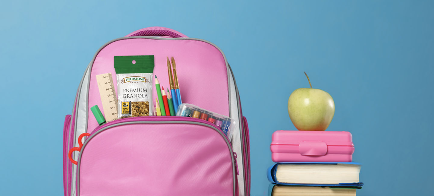 An image of premium granola, and mini doodles in a child's school backpack. A stack of books and art supplies sit next to the backpack with an apple on top. The background of the photo is a bright blue. 
