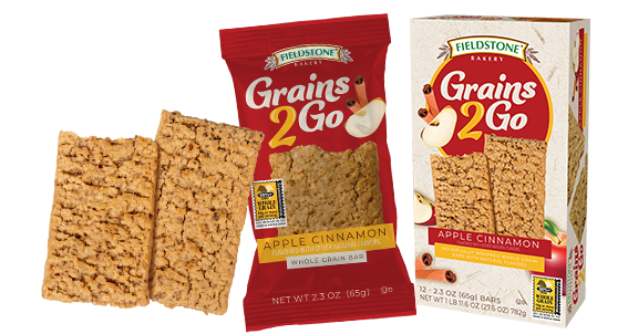 A whole grain apple cinnamon, Grains 2 Go bar in red packaging. An unwrapped chewy granola bar is beside it on the left. A carton of grains 2 go bars are in the back.